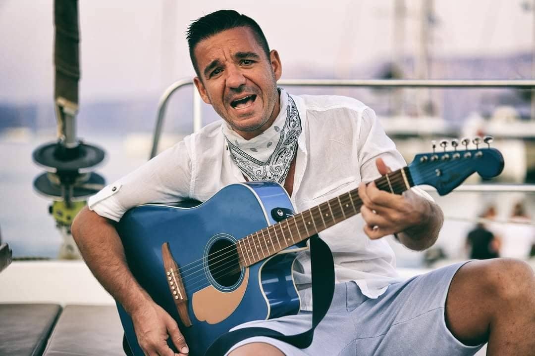 Stan singing and playing a blue acoustic guitar on the deck of a boat, with a harbour behind him.