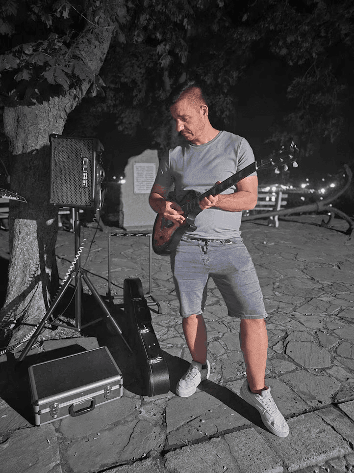 Stan playing an electric guitar at night on a stone-paved square, with an amplifier and a guitar case beside him.
