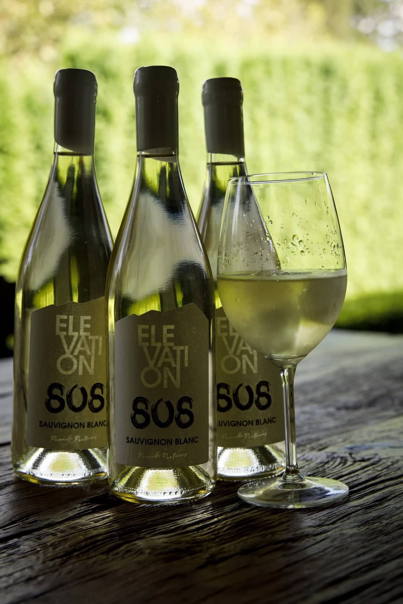 Three bottles of Elevation 808 Sauvignon Blanc and a glass of white wine on a rustic wooden table outdoors.