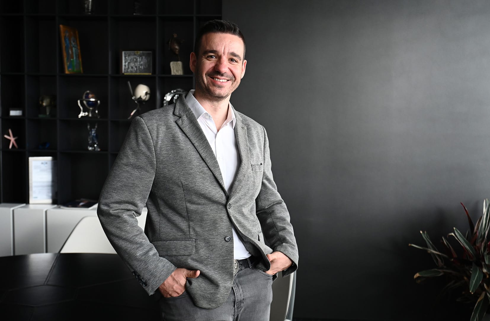 Stanislav Popov smiling, wearing a grey blazer and white shirt, standing in a dark modern office.