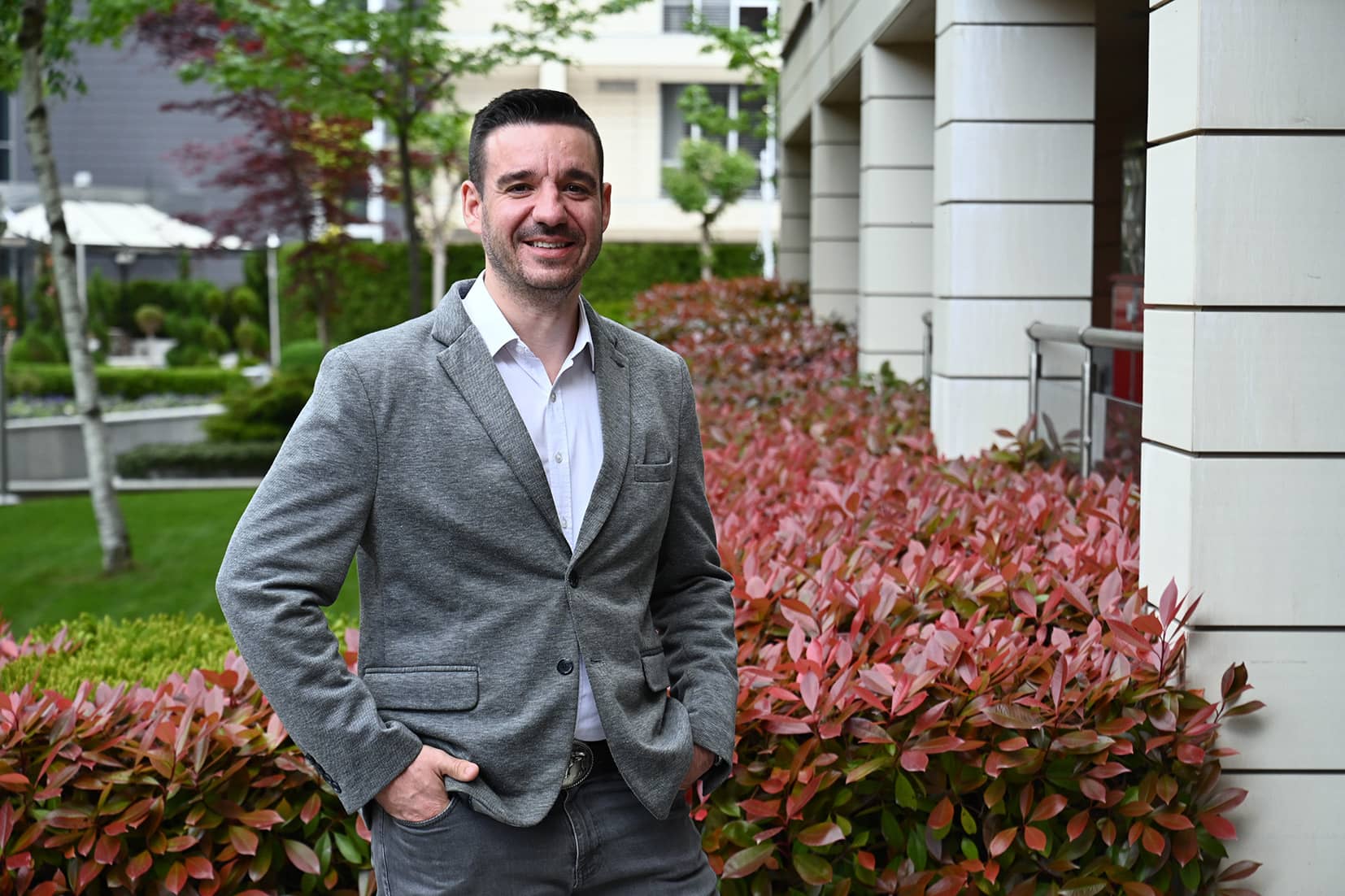 Stanislav Popov in a grey blazer standing outdoors beside red-leafed shrubs and a garden.