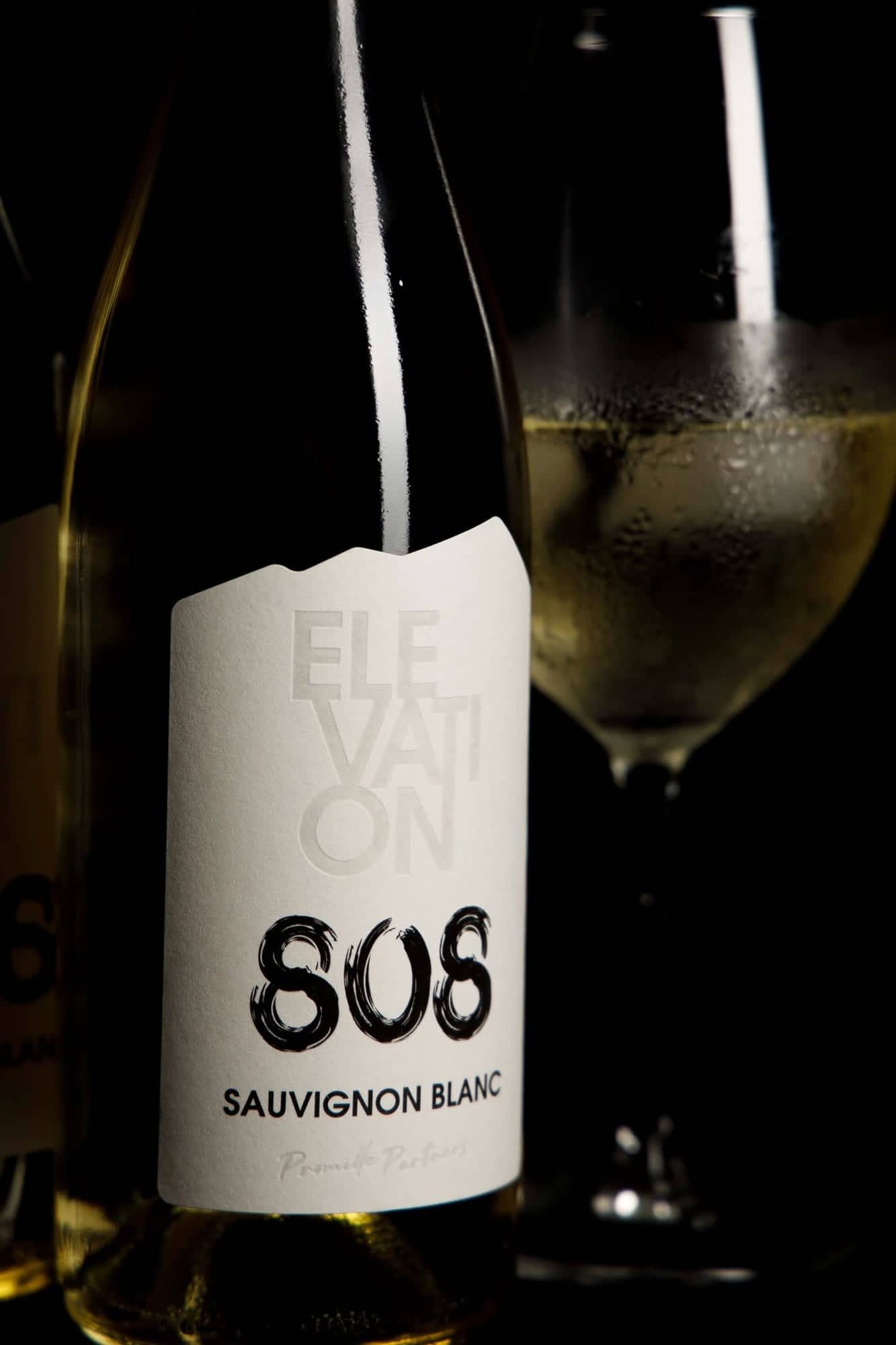 A bottle of Promille Partners Elevation 808 Sauvignon Blanc beside a chilled glass of white wine, on a dark background.