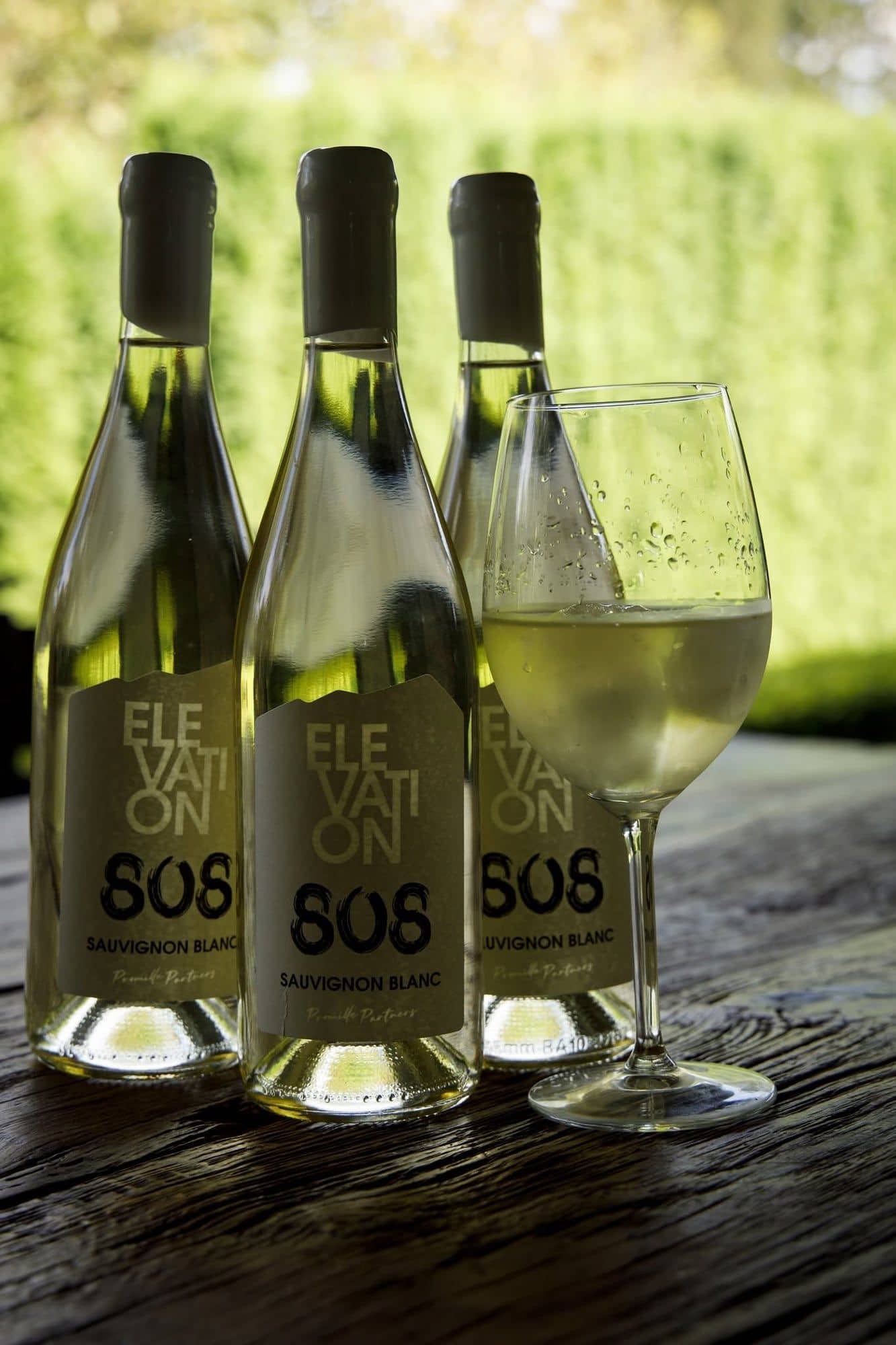 Three bottles of Elevation 808 Sauvignon Blanc and a glass of white wine on a rustic wooden table outdoors.